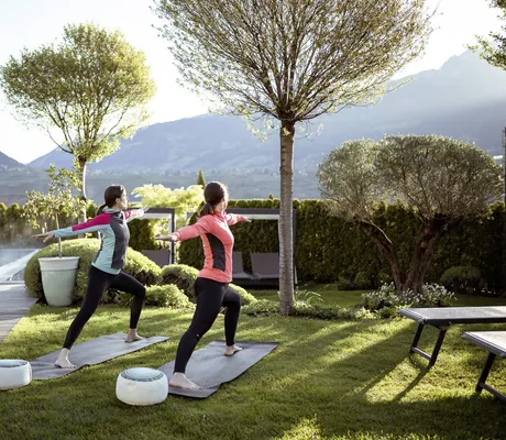 zwei Frauen in Sport-Outfit machen Yoga auf der Wiese neben dem Outdoorpool des La Maiena Meran Resort