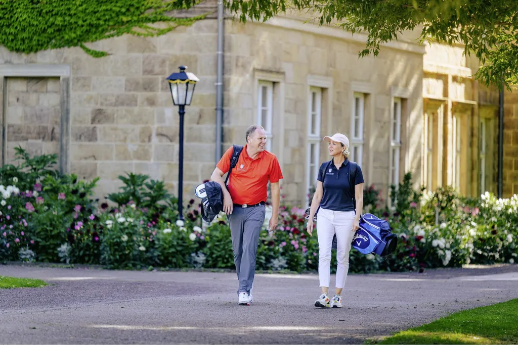 Mann und Frau mit Golf-Equipment laufen über die Anlage des Gräflicher Park Health & Balance Resort