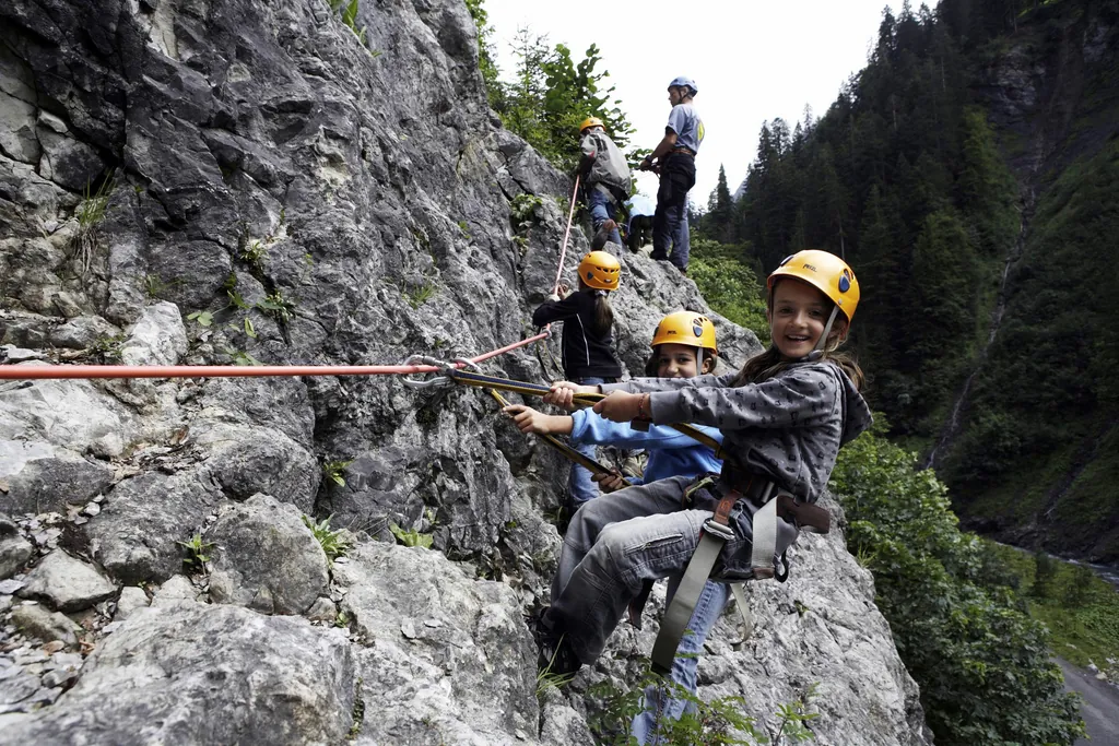 Einige Kinder hängen beim gesicherten Klettern an einer Felswand und lachen in die Kamera