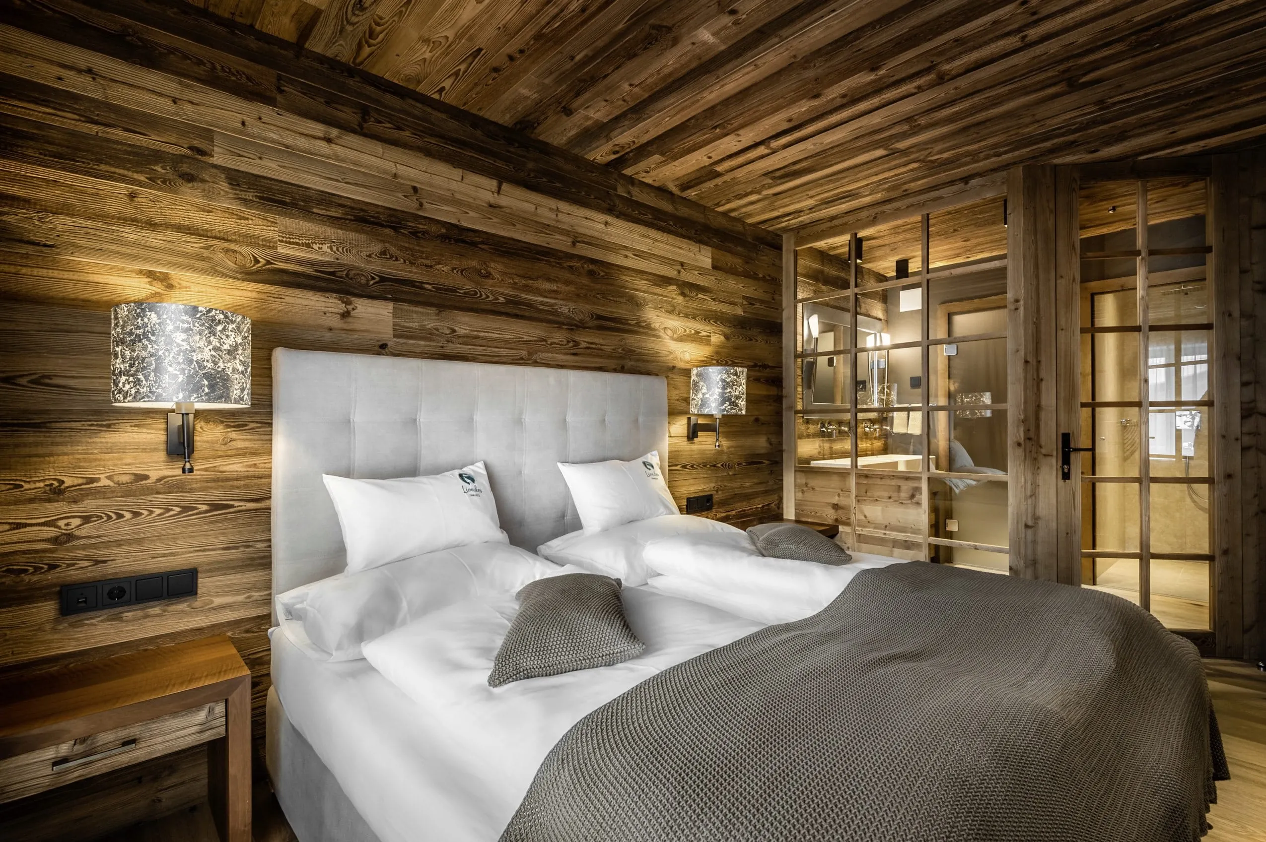 mit dunklem Holz ausgestattetes Schlafzimmer in den Liondes Chalets mit Glast&uuml;r zum Bad