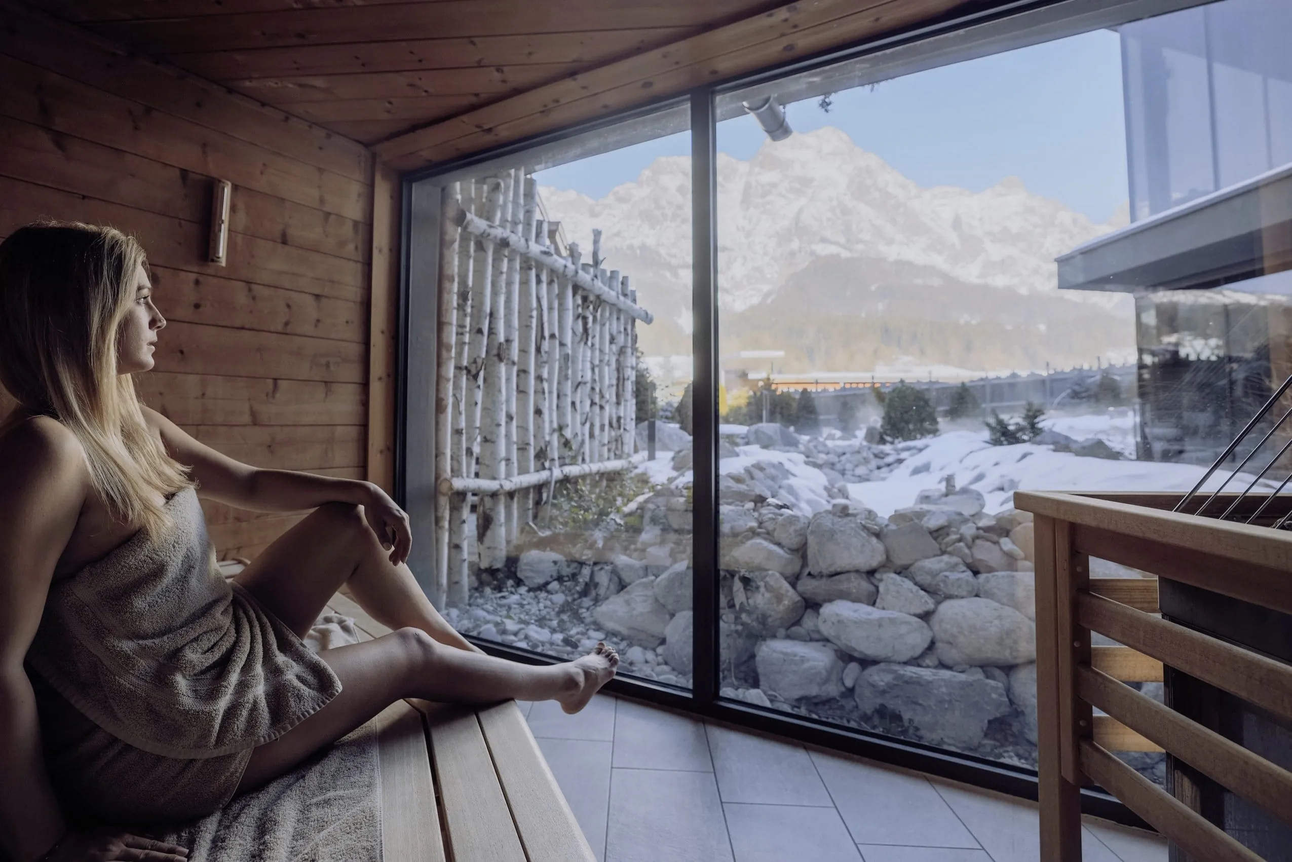 blonde Frau in der Sauna des Good Life Resort Riederalm schaut auf die Berglandschaft