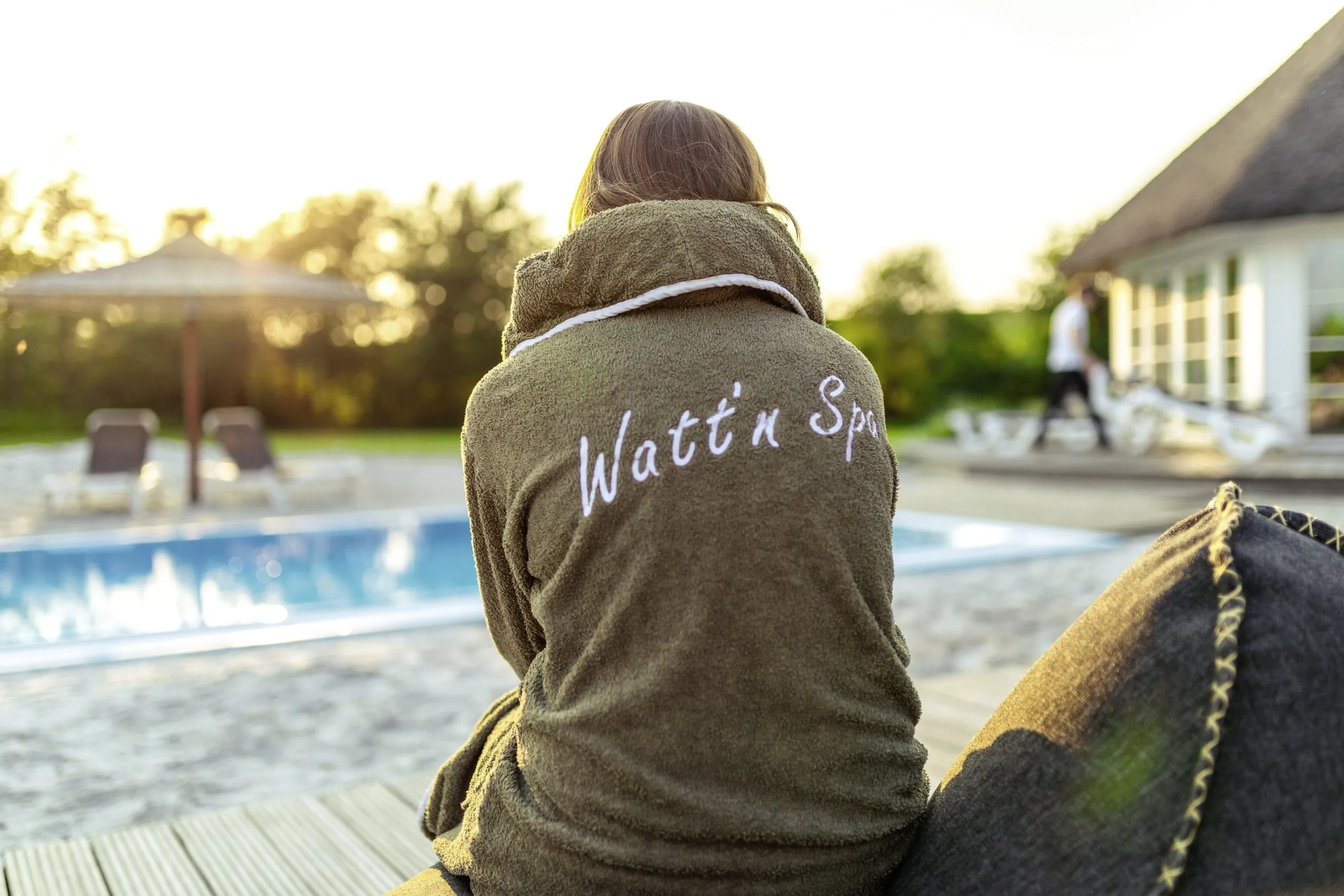 Blick auf den R&uuml;cken einer jungen Frau, die im Bademantel am Outdoorpool des Lundenbergsand Nordsee Hideaway & SPA sitzt