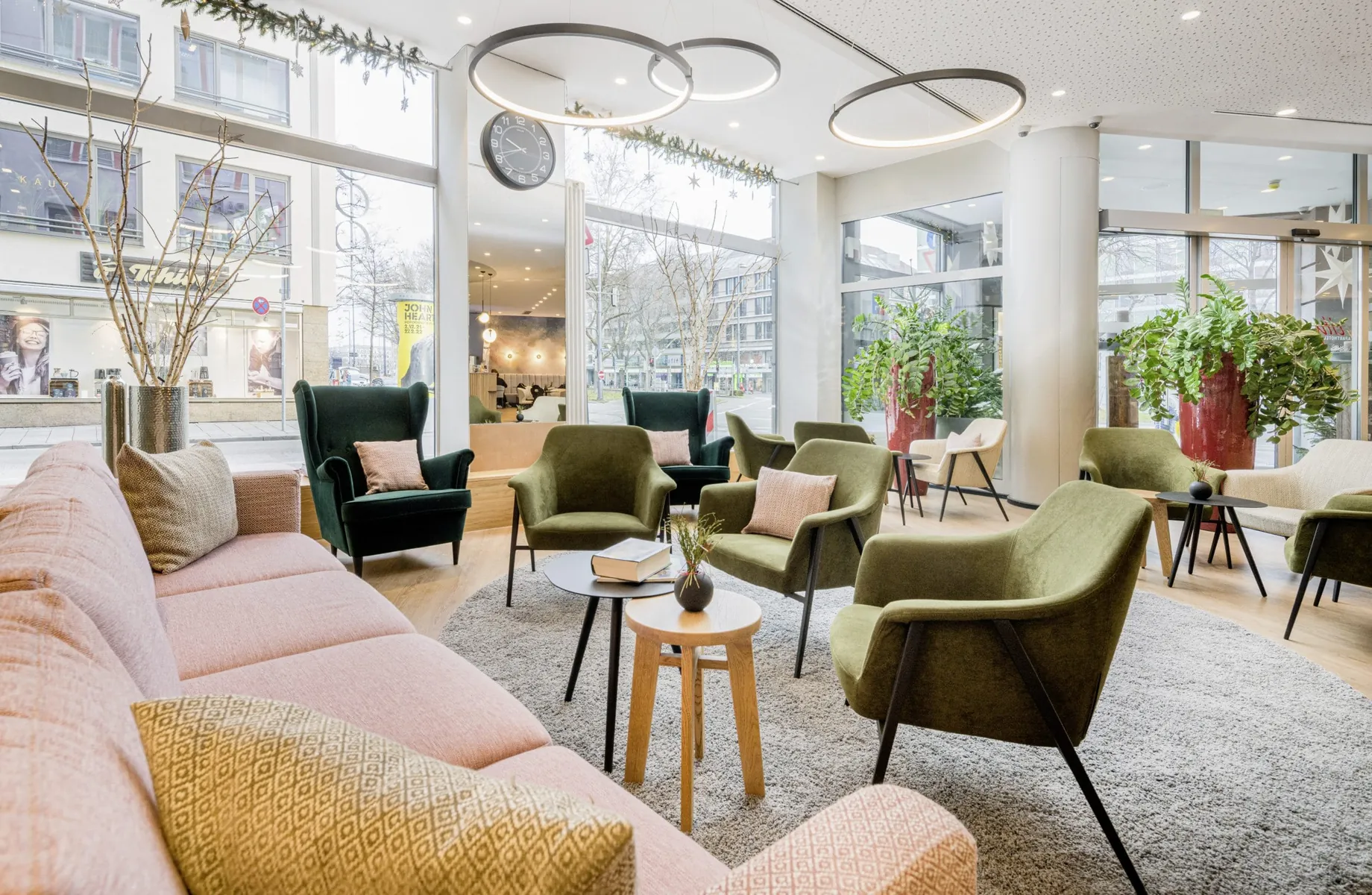 Offene Lobby in skandinavischem Design mit pastellfarbenen Sitzgruppen in grün und rosé mit bodentiefen Fenstern zur Sonnenstraße des KOOS Hotel & Apartments