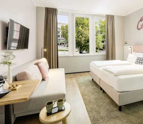 Zimmer in rosé Pastelltönen mit Boxspringbett und darüber stehendem Schriftzug 'Glücksmomente', Sofa und Fensterblick auf Baum mit Häuserfronten im KOOS Hotel & Apartments