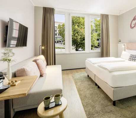 Zimmer in rosé Pastelltönen mit Boxspringbett und darüber stehendem Schriftzug 'Glücksmomente', Sofa und Fensterblick auf Baum mit Häuserfronten im KOOS Hotel & Apartments