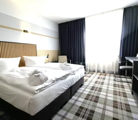 Zimmer mit Boxspringbett mit Holzrückteil, Karo-Teppichboden und schwarzen Vorhängen