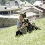 Junge Frau sitzt im Sommer mit Hund in der Wiese, im Hintergrund der Mountain Spring Spa des HUBERTUS Mountain Refugio Allgäu