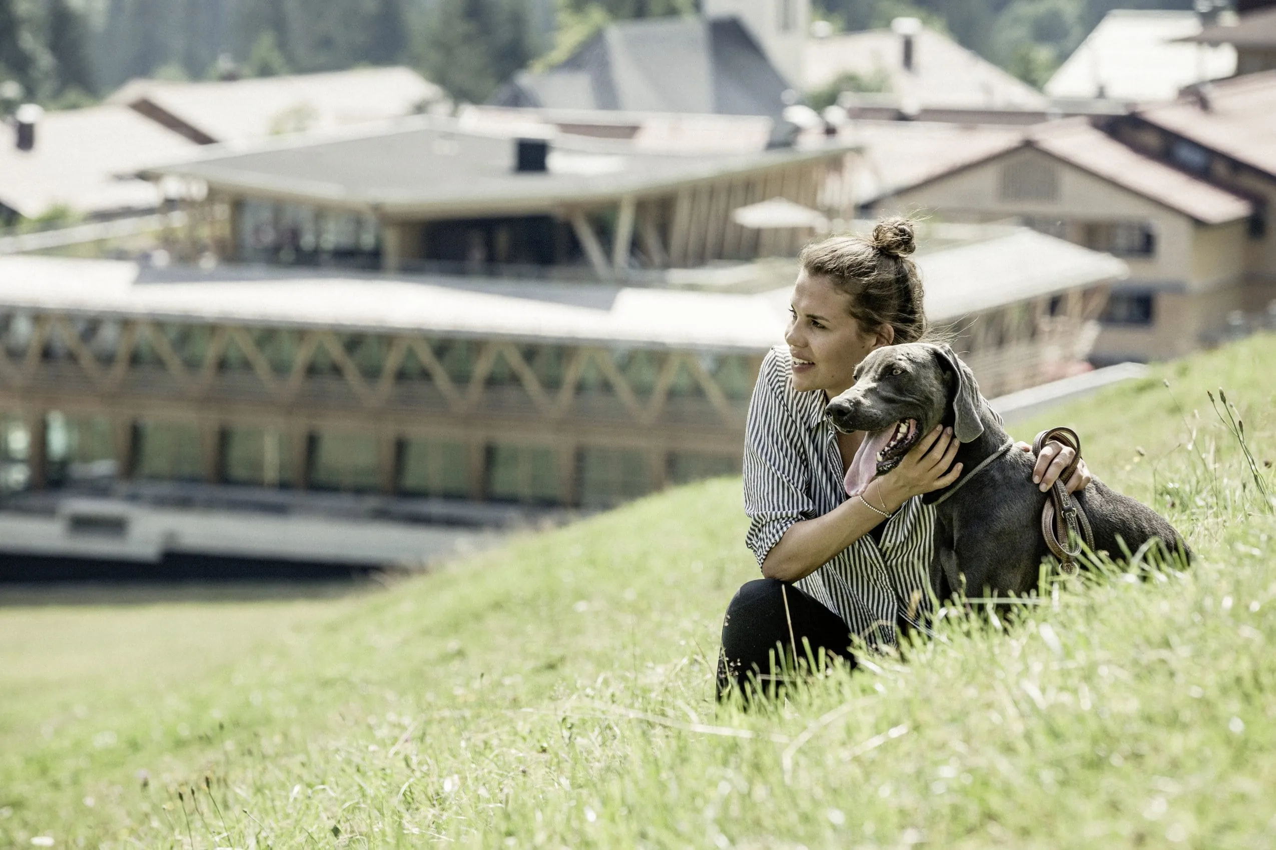 Junge Frau sitzt im Sommer mit Hund in der Wiese, im Hintergrund der Mountain Spring Spa des HUBERTUS Mountain Refugio Allgäu