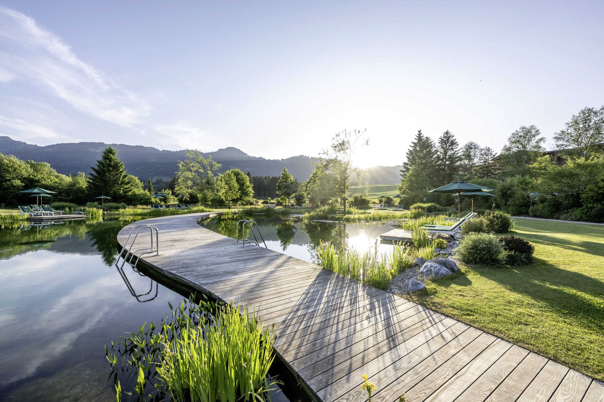 Wunderschön angelegter Naturbadeteich im Wellnesshotel Sonnenalp Resort mit Badesteg und einfallendem Sonnenstrahlen