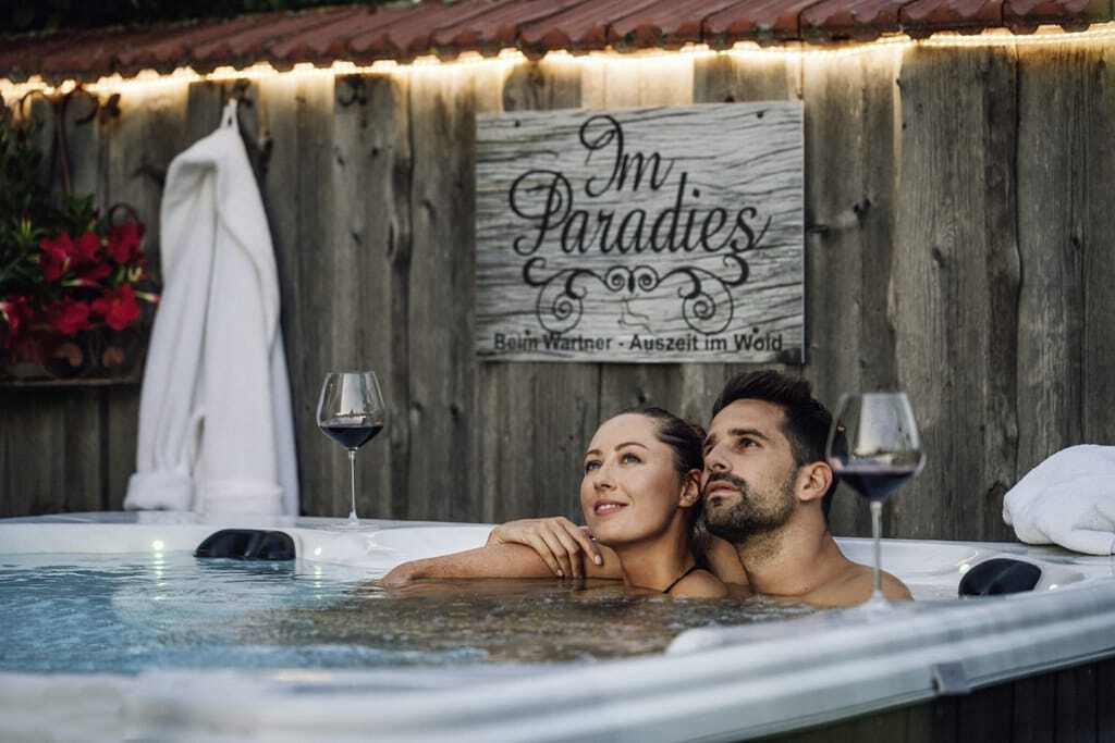 Pärchen sitzt im privaten Whirlpool der Beim Wartner - Chalets & Suiten mit Weingläsern und schaut in den Himmel