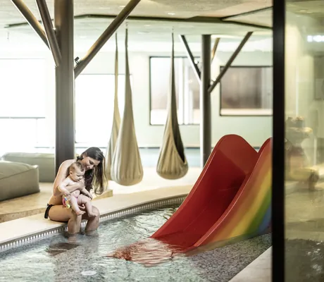 junge Frau sitzt mit Baby auf dem Schoß am Rand des Kinderbeckens mit kleiner Wasserrutsche im Falkensteiner Family Resort Lido