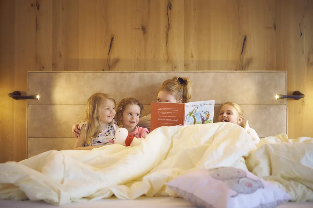 blonde Frau sitzt mit drei Mädchen im Bauernhof Hotel Oberschwarzach im Bett und liest ein Buch vor