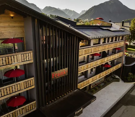 Fassade des Hotel Franks mit einigen Balkonen, roten Sonnenschirmen und Bergen im Hintergrund