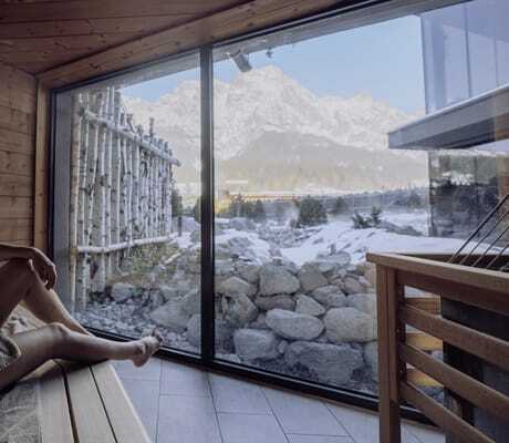 blonde Frau sitzt mit Handtuch in der Sauna der Riederalm und blickt auf die Winterlandschaft