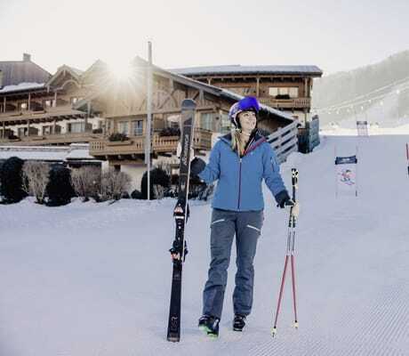 blonde Frau mit blauem Skioutfit trägt ihre Skier auf der Piste vor der Riederalm