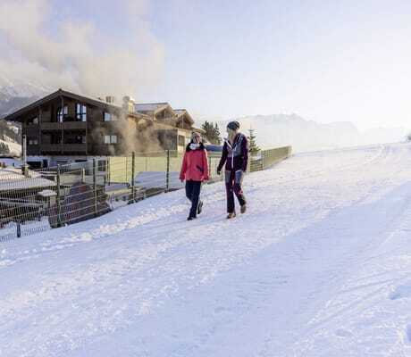 zwei Frauen in Winteroutfit wandern im Schnee an der Riederalm vorbei