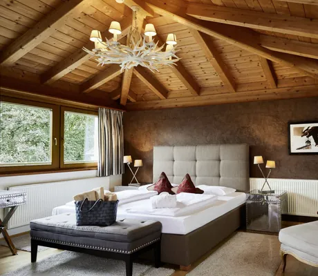 Blick in die elegante Schwarzbrunn Suite des Hotel Schwarzbrunn im modernen Landhausstil mit Doppelbett, Kronleuchter mit kleinen Lampenschirmen und hellgrauem Sessel