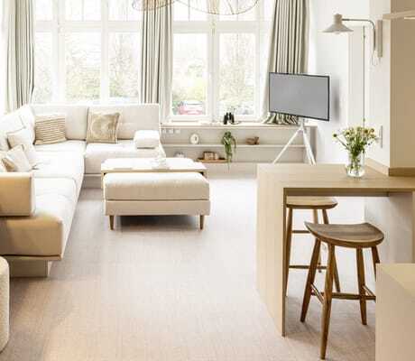 lichtdurchfluteter Wohnbereich einer der Suiten in der Villa Astoria mit großen Fenstern, großem beigefarbenem Sofa, Flachbild-Fernseher und zwei Sitzhockern aus Holz