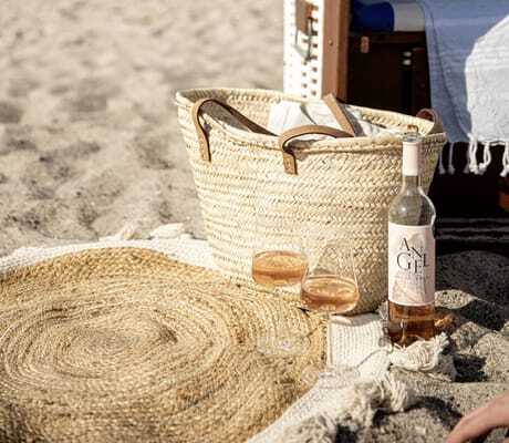 kleine, runde Strandmatte im Boho-Stil mit zwei gefüllten Weingläsern, Korbtasche und Strandkorb in der Nähe der Villa Astoria