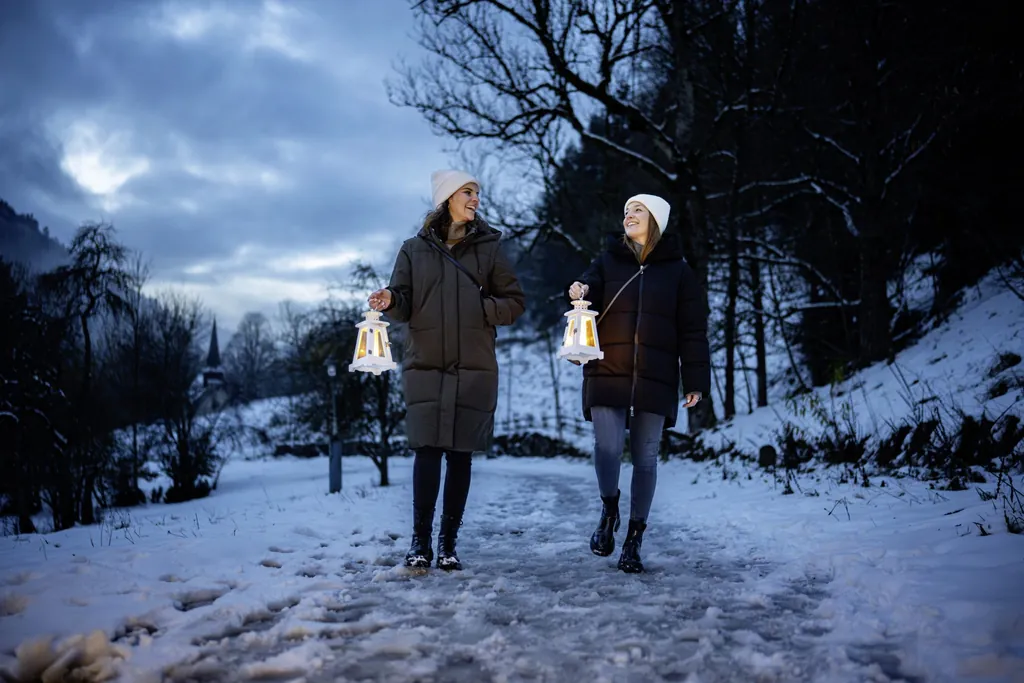Zwei junge Frauen mit Wollm&uuml;tzen und Wintermantel laufen bei Abendd&auml;mmerung auf einem verschneiten Weg mit Laternen in der Hand und l&auml;cheln sich an