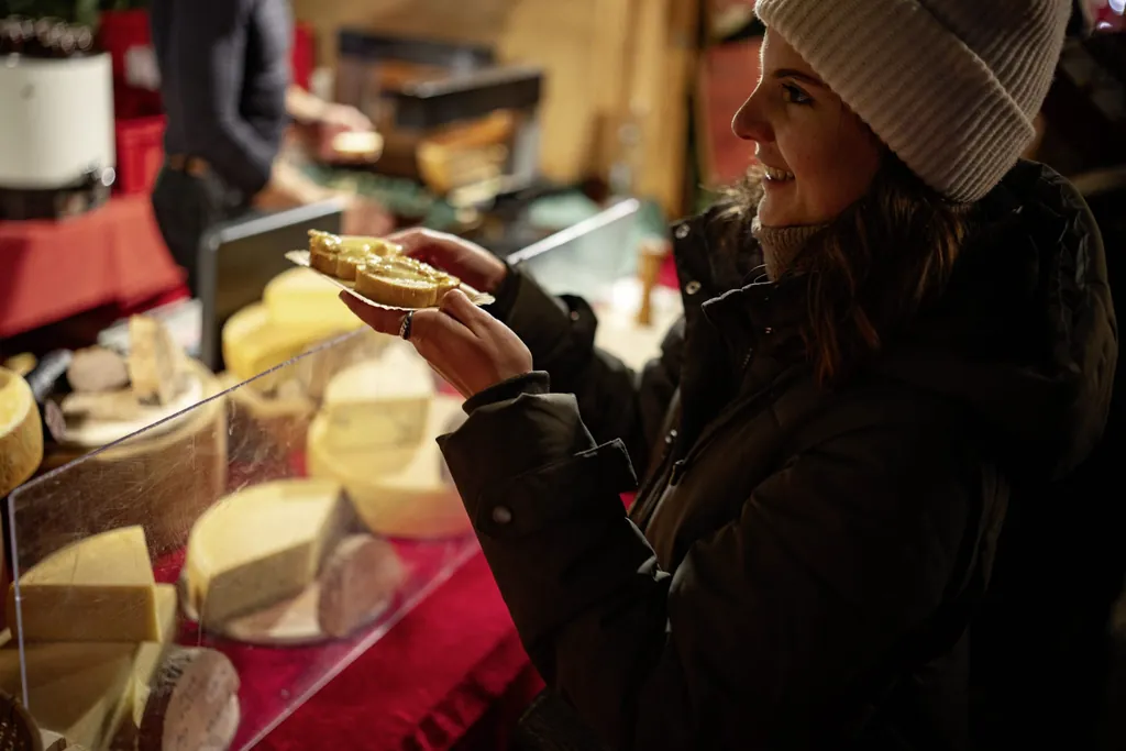 Junge Frau mit Wollm&uuml;tze nimmt zwei Raclettebrote an einem Weihnachtsmarktstand entgegen