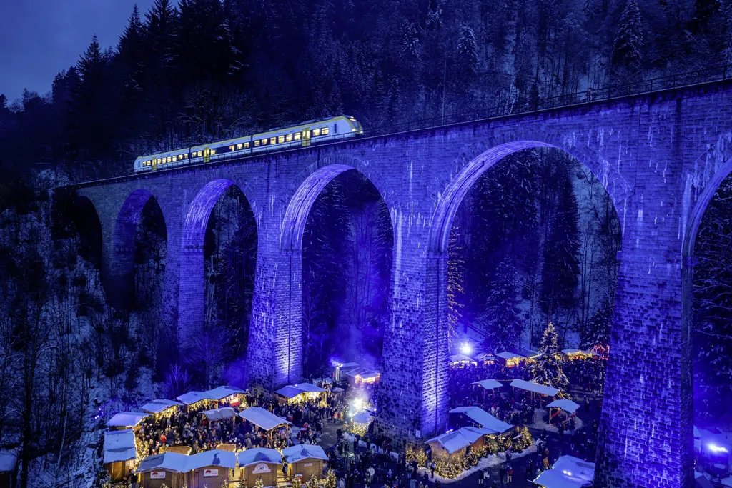Ein beleuchteter Zug f&auml;hrt in der Abendd&auml;mmerung &uuml;ber die Viaduktb&ouml;gen der Ravennschlucht, in der ein Weihnachtsmarkt stattfindet