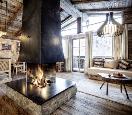 Wohnbereich in Holz- und Steinoptik in einem der Chalets des Bergdorf Prechtlgut mit verglastem Kamin in der Mitte, Sitzbank und Küche