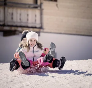 Kinder rodeln lachend im Schnee vor einem Holzhaus des Feuerstein Nature Family Resort