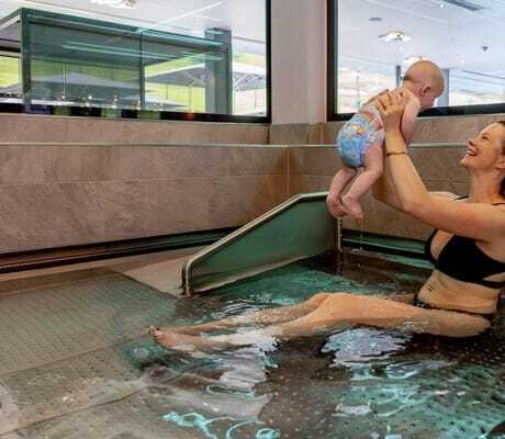 Frau mit Pferdeschwanz hebt im Babypool der Riederalm ein Baby in die Luft
