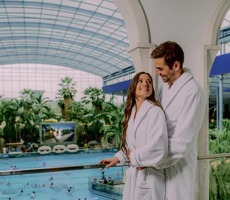 Paar in weißen Bademänteln im Hotel Victory Therme Erding lächelt sich an mit großem Pool und Palmen im Hintergrund