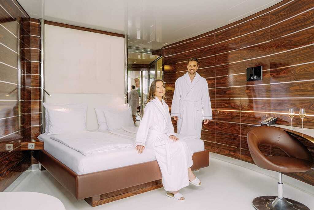 junges paar in Bademänteln in einem der braun-weiß eingerichteten Zimmer des Hotel Victory Therme Erding mit braunem Lederstuhl