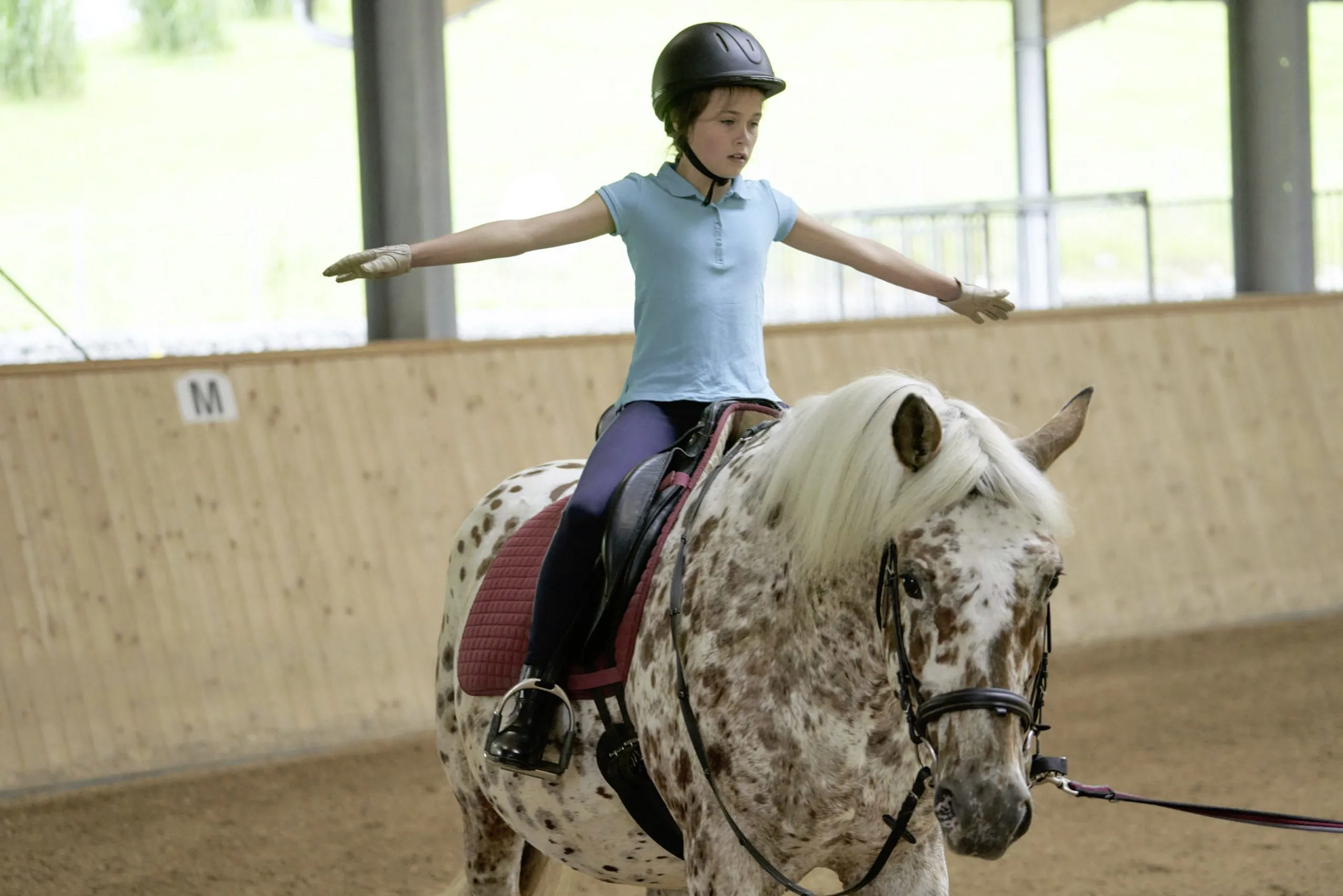 M&auml;dchen sitzt auf Pferd mit ausgestreckten Armen in Reithalle des Familotel Schreinerhof
