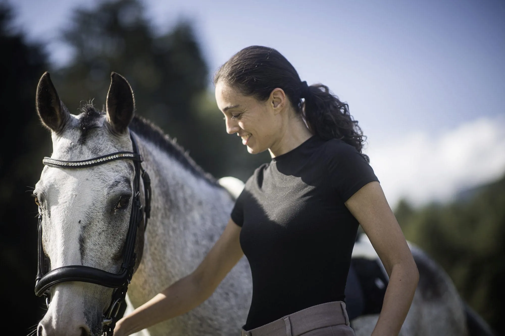 junge dunkelhaarige Frau l&auml;chelt wei&szlig;-graues Pferd des Hotel Winkler an, das neben ihr steht