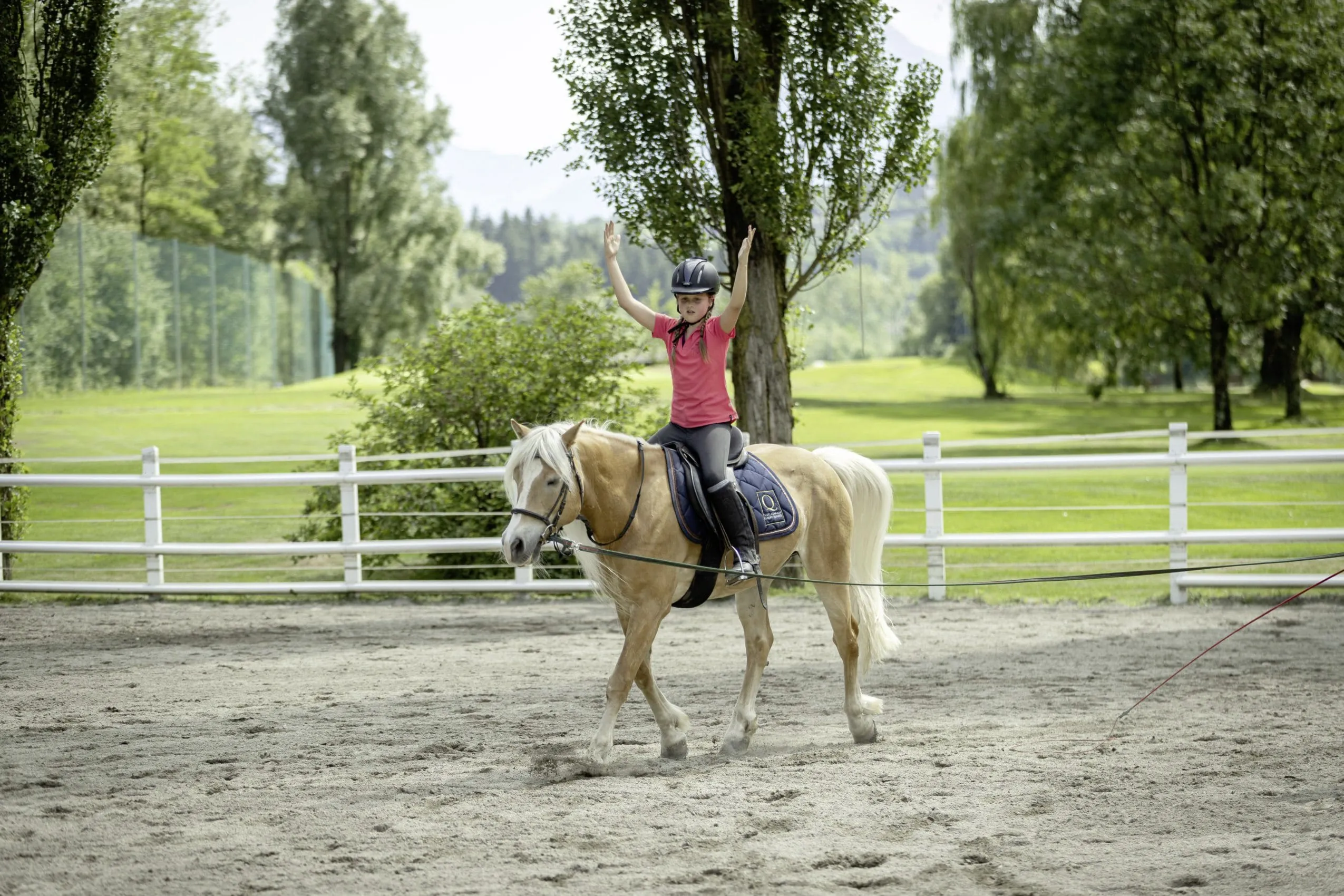 junges M&auml;dchen auf Pferd auf dem Reitplatz des Quellenhof Luxury Resort Passeier h&auml;lt die Arme nach oben