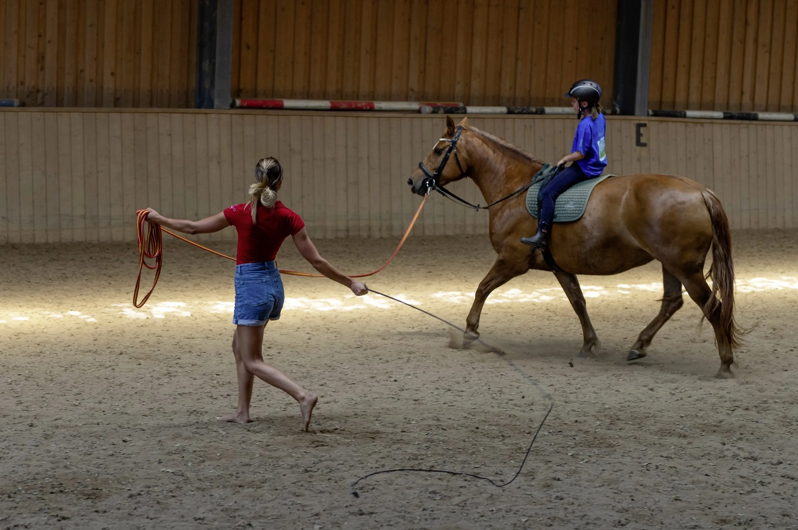 Reitlehrerin leitet Pferd mit Kind mit Seil und Peitsche in der Reithalle des Ulrichshof an