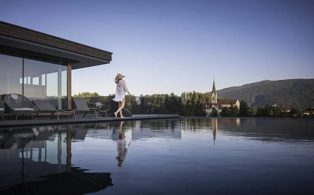 Frau mit Sonnenhut und leichtem Sommerkleid geht am Rand des Panorama-Infinity-Rooftop-Sky-Pool im active lifestyle hotel Terentnerhof