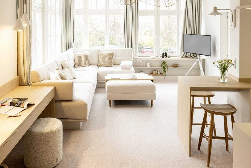 lichtdurchfluteter Wohnbereich einer der Suiten in der Villa Astoria mit großen Fenstern, großem beigefarbenem Sofa, Flachbild-Fernseher und zwei Sitzhockern aus Holz