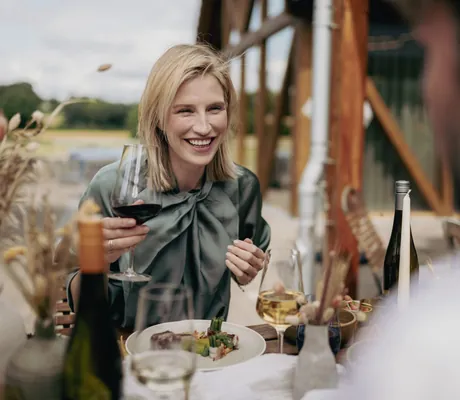 blonde Frau mit grüner Bluse und Rotweinglas sitzt lachend am gedeckten Tisch in Jordan's Untermühle
