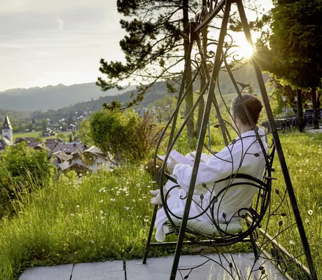 Frau in Bademantel sitzt mit Buch auf der Hollywoodschaukel des Hotel Prinz-Luitpold-Bad und blickt in die Natur