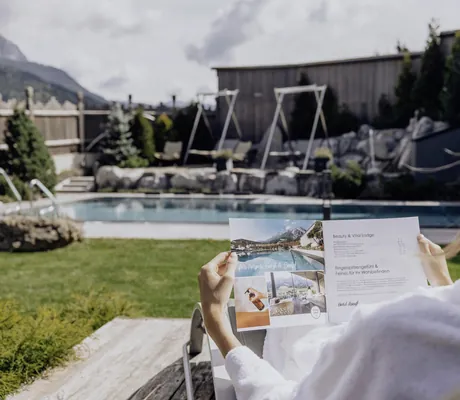 Frau liegt auf einer Liege im Garten der Riederalm und liest die Spa-Broschüre