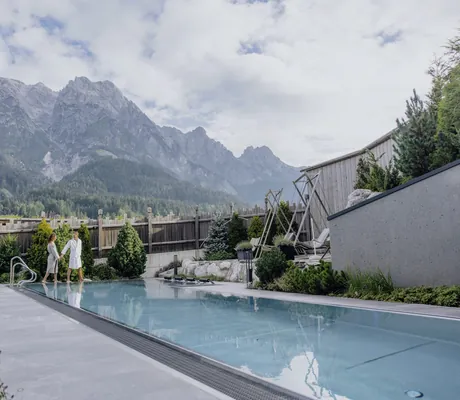 Paar in weißem Bademantel am Beckenrand des Thermalwasserpools der Riederalm vor der Bergkulisse