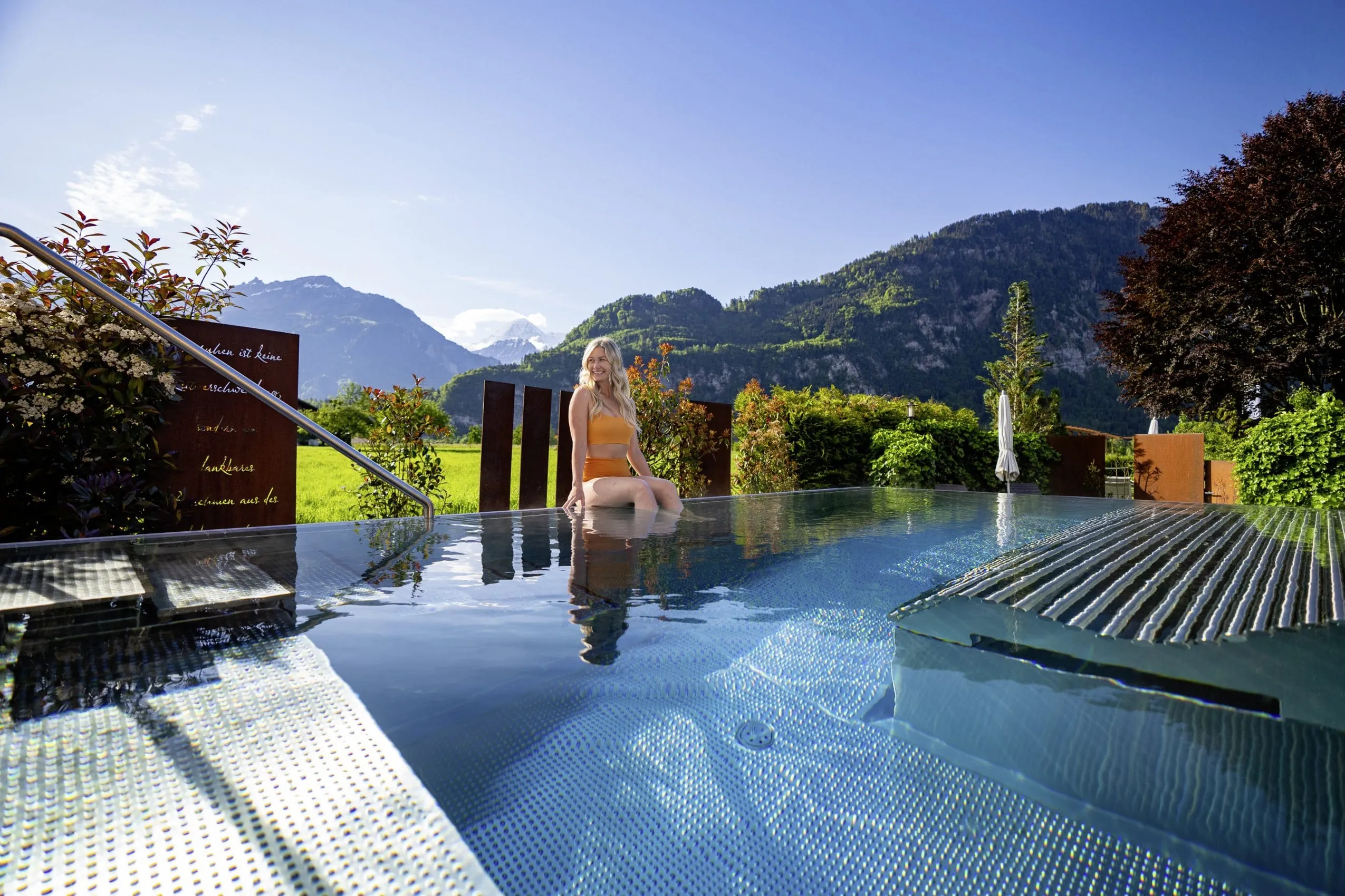 junge blonde Frau in orangefarbenem Bikini sitzt am Au&szlig;enpool des Salzano Hotel-Spa-Restaurant mit Bergen im Hintergrund