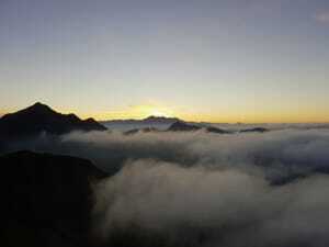 Blick auf Berggipfel bei Sonnenaufgang mit Wolkenmeer darunter und gelb-orangenem Sonnenlicht am Himmel