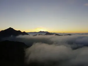Blick auf Berggipfel bei Sonnenaufgang mit Wolkenmeer darunter und gelb-orangenem Sonnenlicht am Himmel