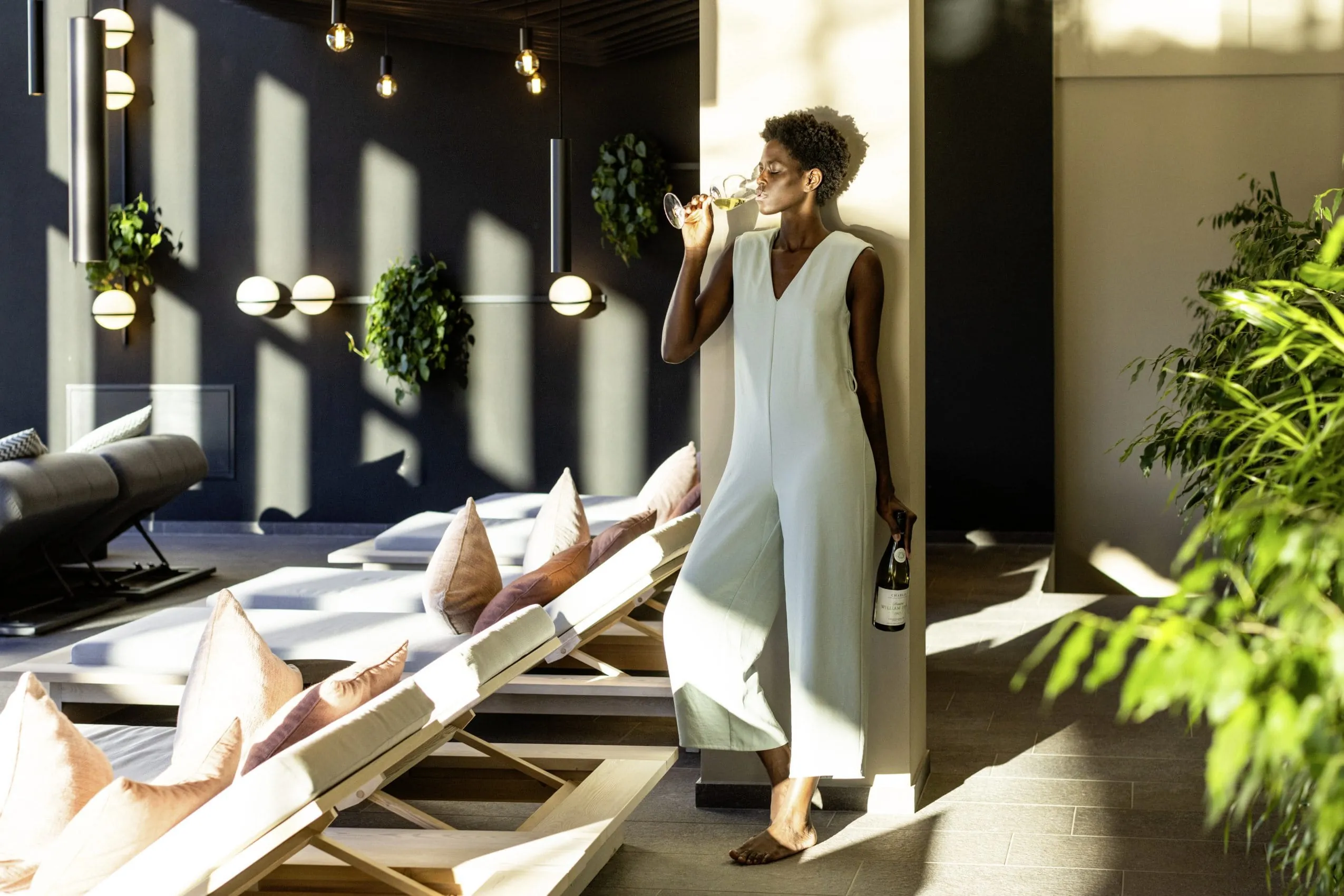 dunkelhäutige Frau in weißem Jumpsuite trinkt neben den Ruheliegen im Hotel Weisses Kreuz ein Glas Wein