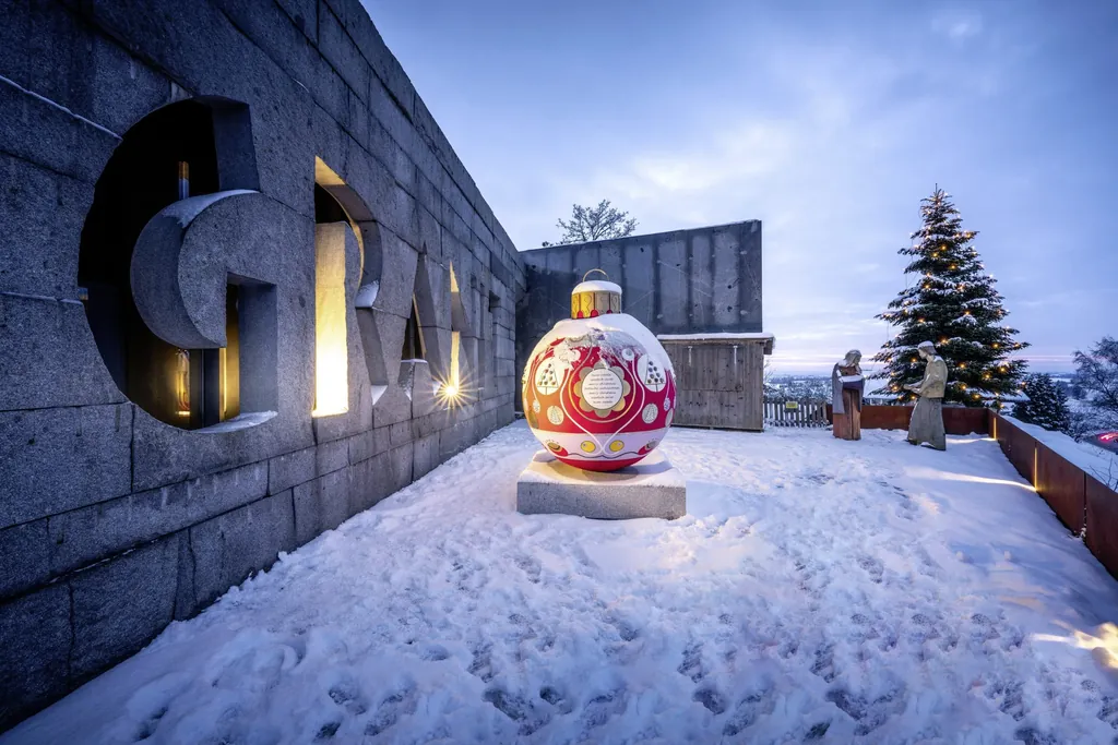 Gro&szlig;e Christbaumkugel im Schnee vor dem Granitzentrum Hauzenberg mit zwei Krippenfiguren und gro&szlig;em, beleuchteten Tannenbaum im Hintergrund