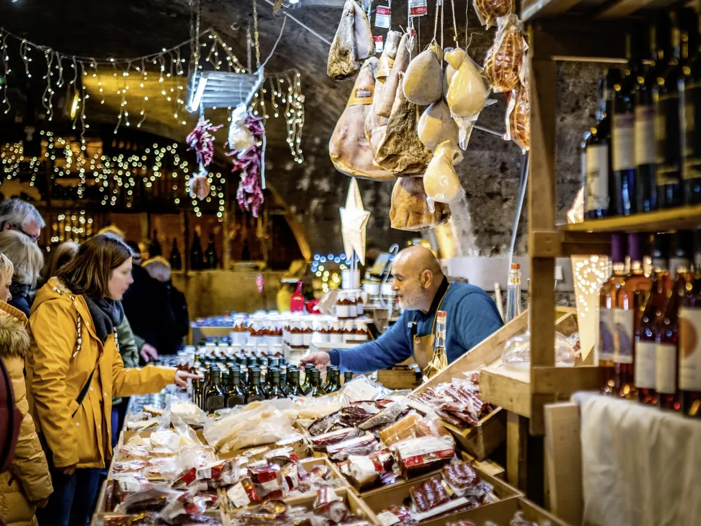 Frau in gelber Jacke steht an einem Marktstand mit Lebensmitteln, Schinken und Wein auf dem Mosel-Wein-Nachts-Markt in einem Weinkeller
