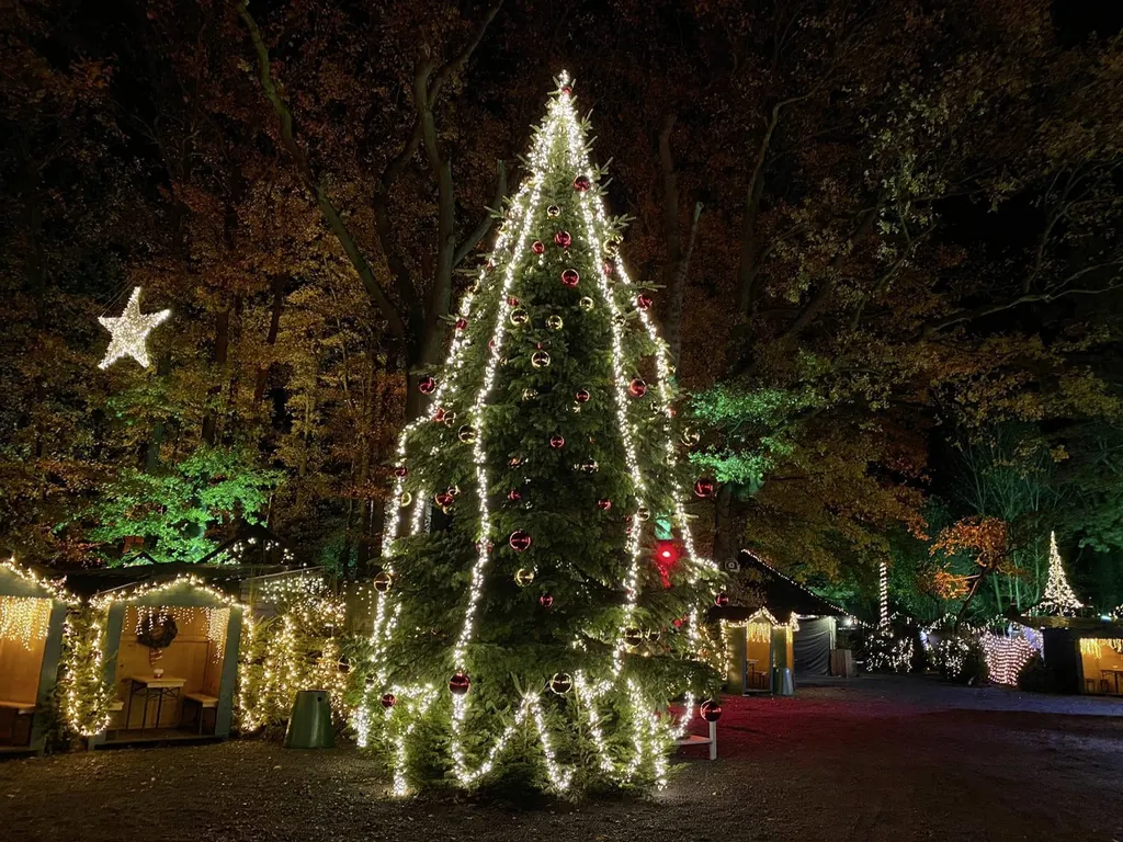 Gro&szlig;er mit Kugeln und mit Lichterketten dekorierter Tannenbaum und beleuchtete Weihnachtsmarktst&auml;nde im Hintergrund