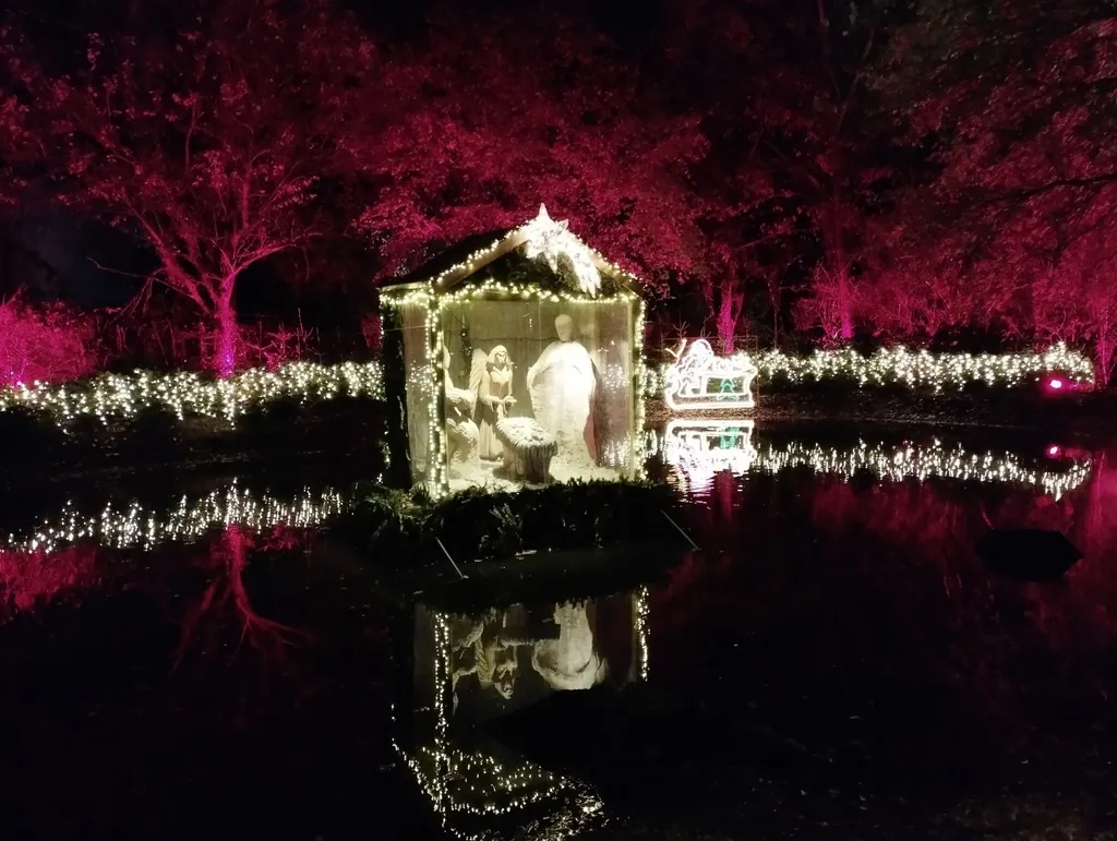 Kleine Krippe mit Holzfiguren auf einem See, geschm&uuml;ckt mit Lichterketten und einem leuchtenden Weihnachtsstern und weiteren Lichtilluminationen im Hintergrund