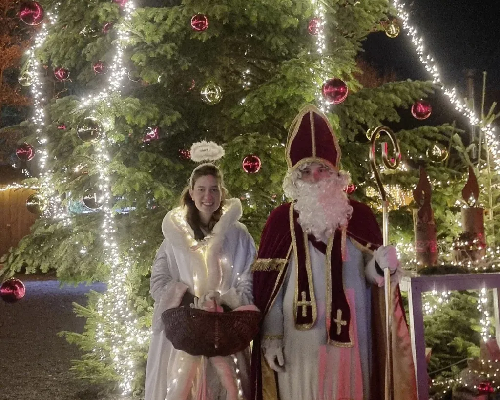 Junge Frau in Engelskost&uuml;m mit Heiligenschein und junger Mann in Nikolauskost&uuml;m stehen vor einem beleuchteten und mit Kugeln geschm&uuml;cktem Tannenbaum